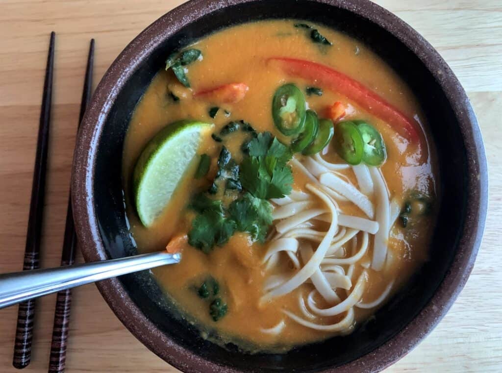vegan creamy curried red lentil noodle soup with lime jalapeno cilantro and bell pepper in a brown ceramic bowl with a spoon and chopsticks