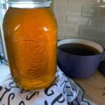 vegetable broth in a glass mason jar on a white towel with black words a purple saucepan and grey tile background