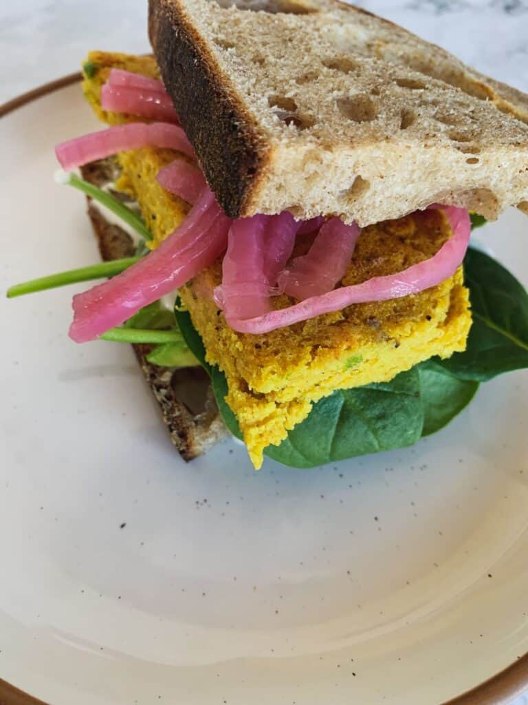 chickpea egg sandwich on sourdough bread with pickled onion and spinach on white plate