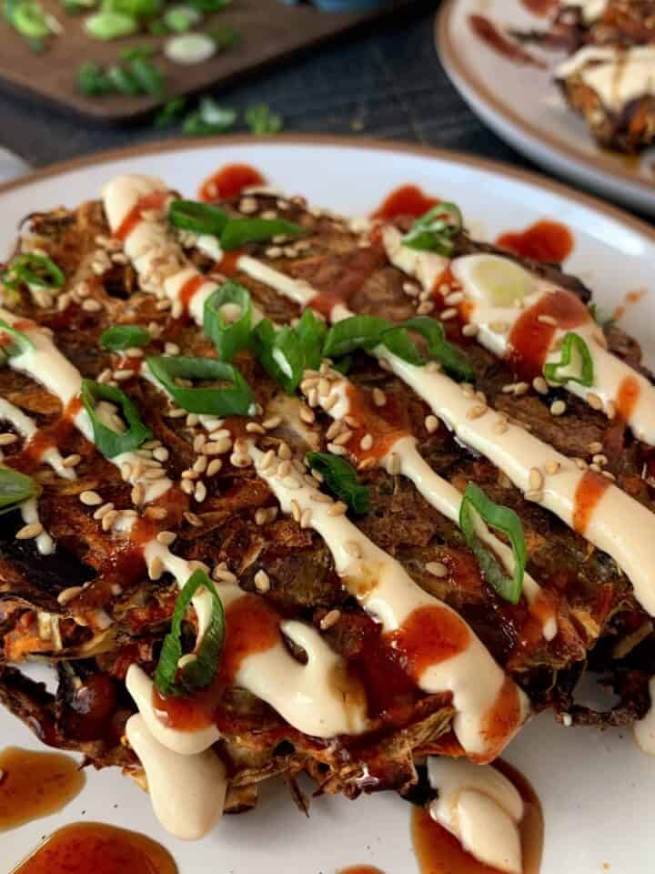 baked vegan okonomiyaki on a white plate topped with spicy mayo, sriracha, green onions and sesame seeds