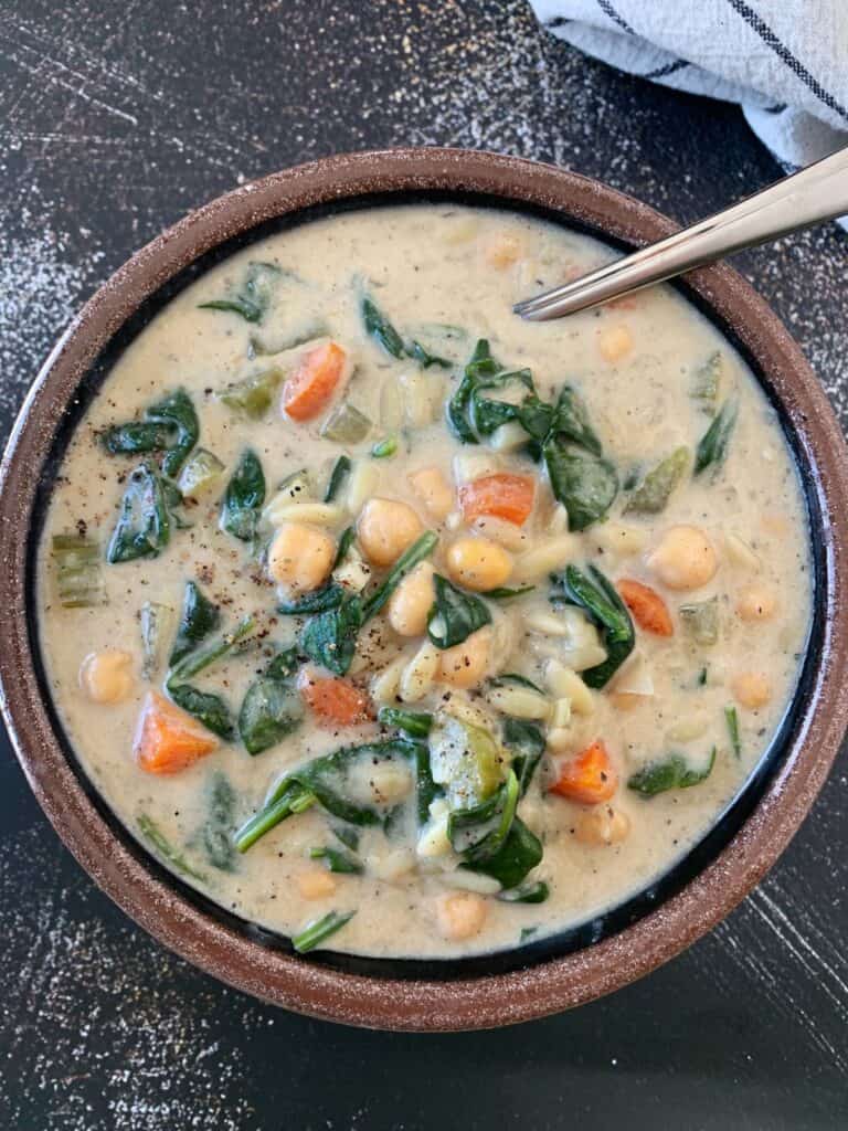 Creamy chickpea orzo soup recipe in a brown ceramic bowl with pepper on top and a silver spoon