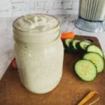 creamy vegan dill salad dressing in a mason jar on a brown cutting board with cucumbers and carrot sticks
