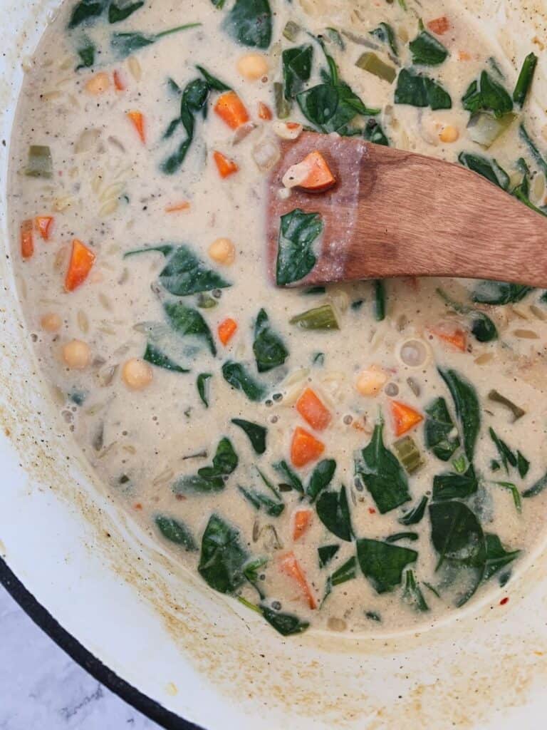 close up view of creamy orzo vegetable soup in a pot with a wooden utensil