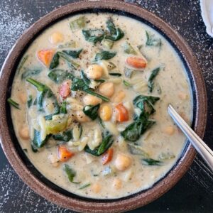 vegan chickpea orzo soup in a brown bowl with a spoon and dark background