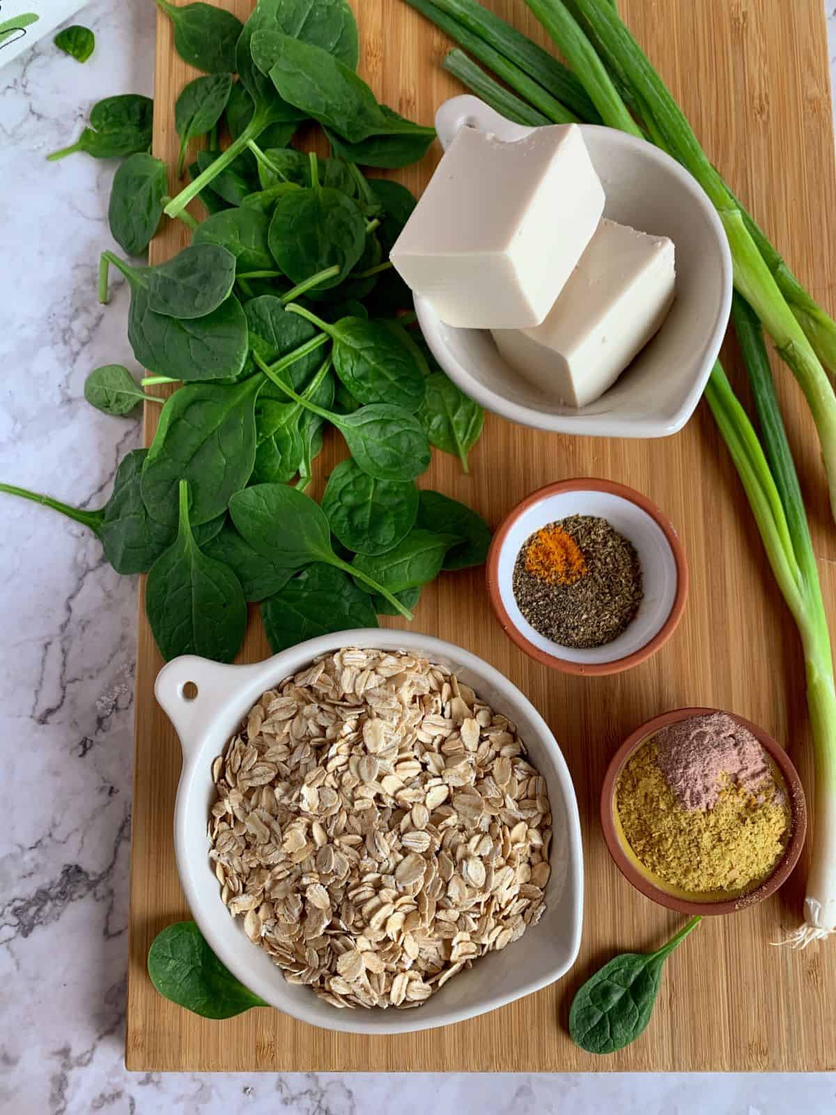 ingredients for savory oatmeal, oats, spinach, spices, tofu and green onions, laid out on a wooden board.