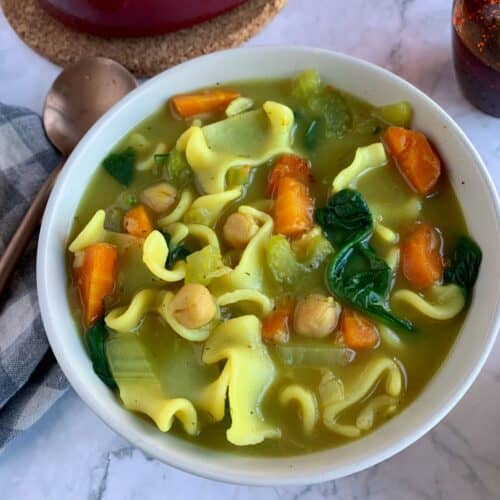 Vegan chickpea noodle soup with spinach in a white bowl with gold spoon.