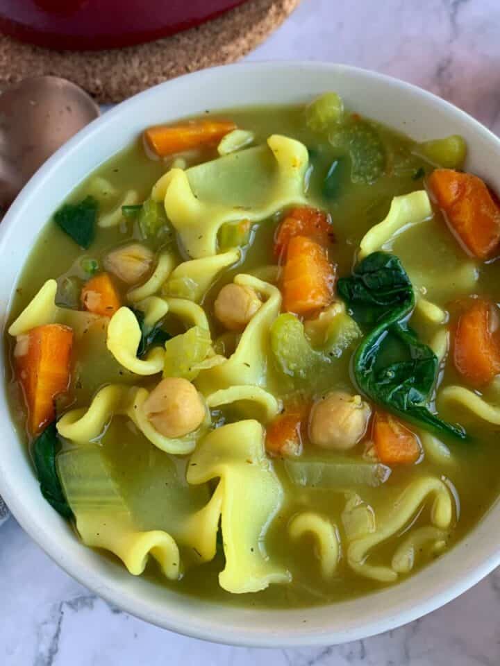 Vegan chickpea noodle soup with spinach in a white bowl with gold spoon.