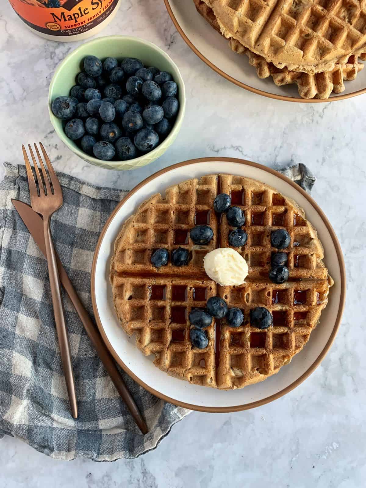 Oatmeal waffle on a white plate with blueberries, maple syrup and vegan butter.